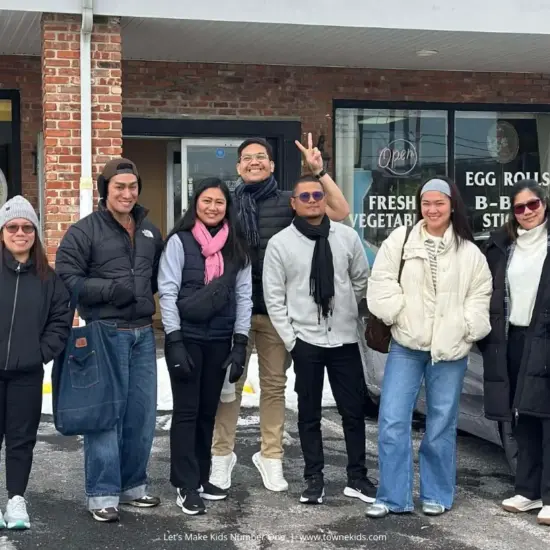 Seven adults in winter clothing smiling and posing together outside a storefront.