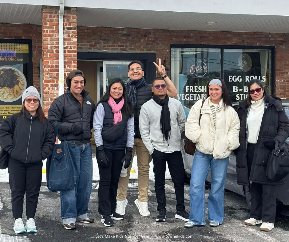 Seven adults in winter clothing smiling and posing together outside a storefront.